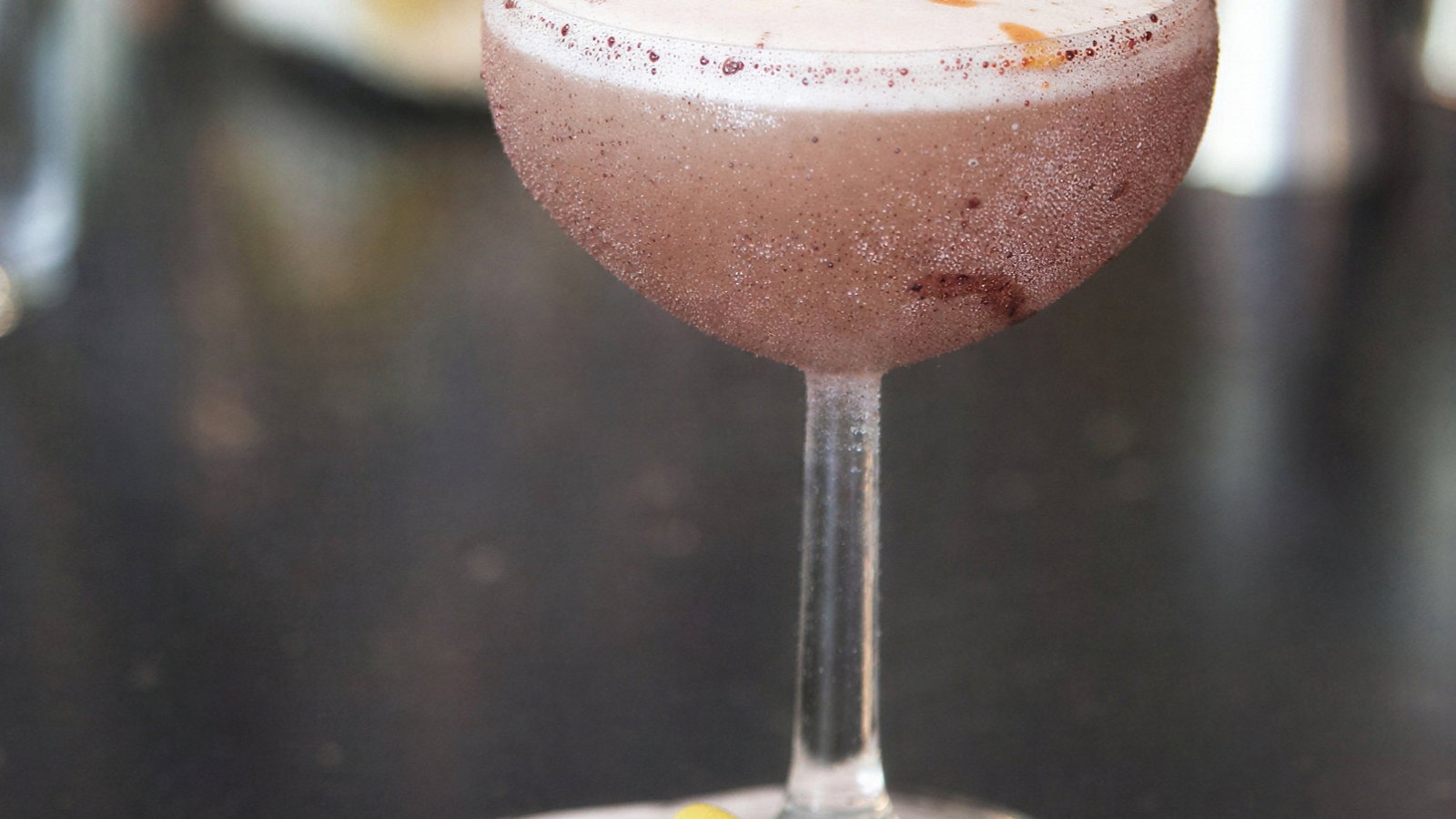 A frothy cocktail in a coupe glass rests on a white coaster with a lemon twist garnish. Blurred background, dark surface.