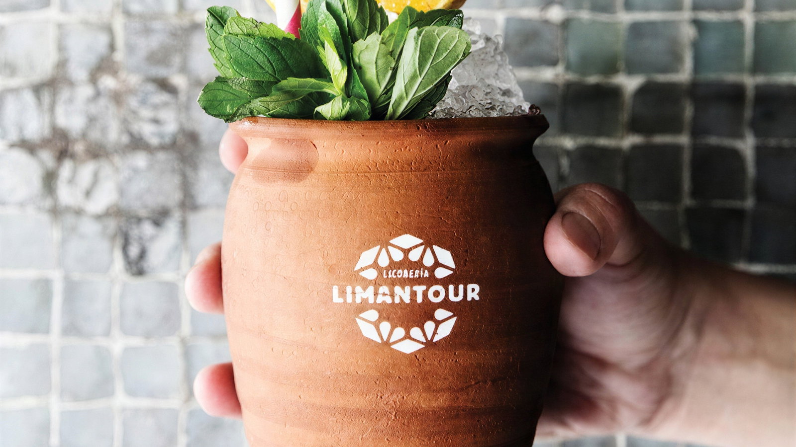 Hand holding a clay cup with mint leaves, ice, an orange slice, and a straw. "Licorería Limantour" text on cup. Tiled gray background.