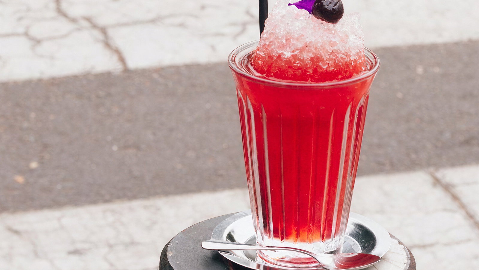 Red iced drink with a straw and fruit garnish on a pedestal. Set on a street with a crosswalk in the background, creating a refreshing mood.