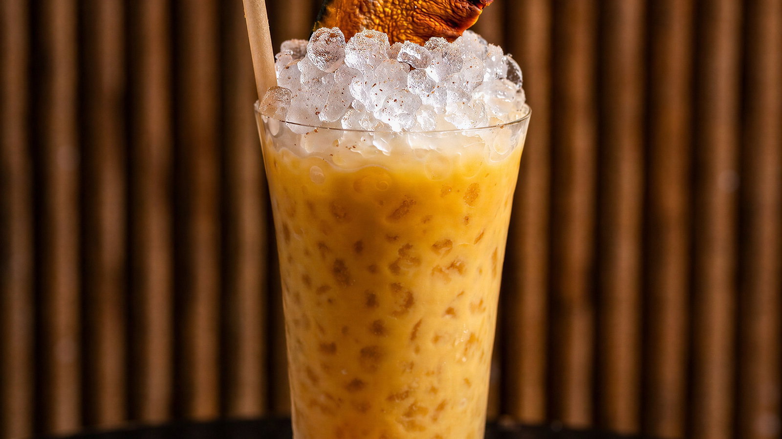 Orange drink with ice, garnished with a dried citrus slice and straw, on a multi-colored coaster. Background features wooden slats.