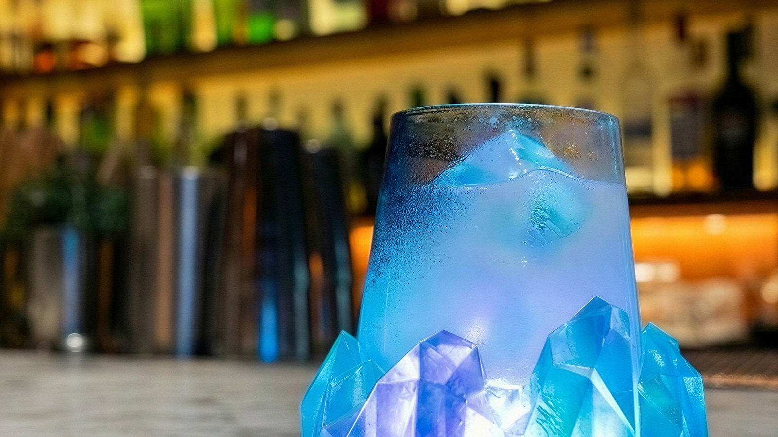 A glowing blue cocktail in a crystal-like glass sits on a marble bar counter, with blurred shelves of bottles in the warm-lit background.