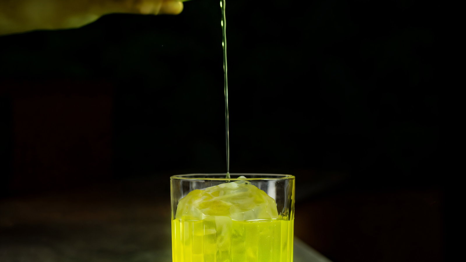 Hand pours yellow liquid into glass with large ice sphere on wooden coaster, set on a green marble table, against a dark background.