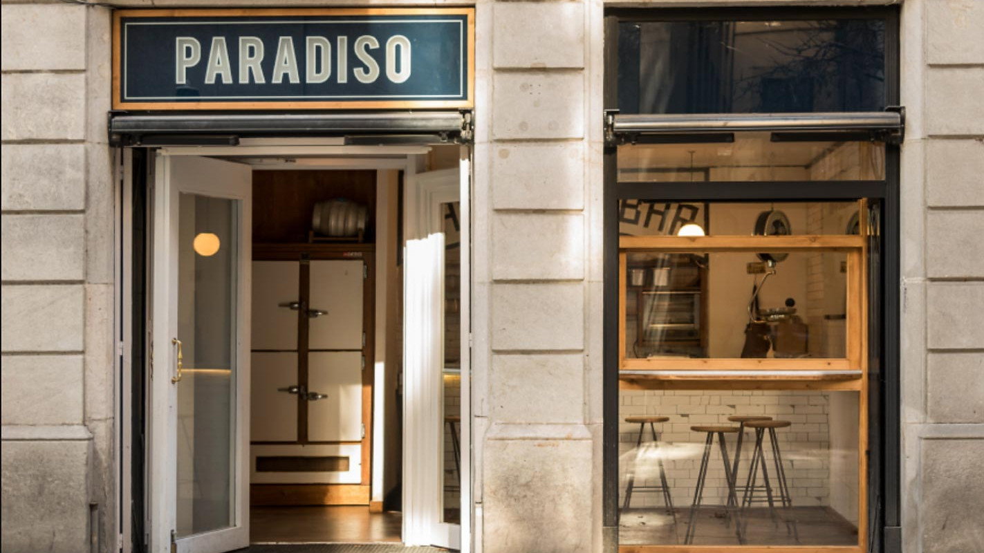 Facade of a building with an open door. Sign reads "PARADISO." Inside view shows bar stools and shelves. Neutral-toned stone exterior.