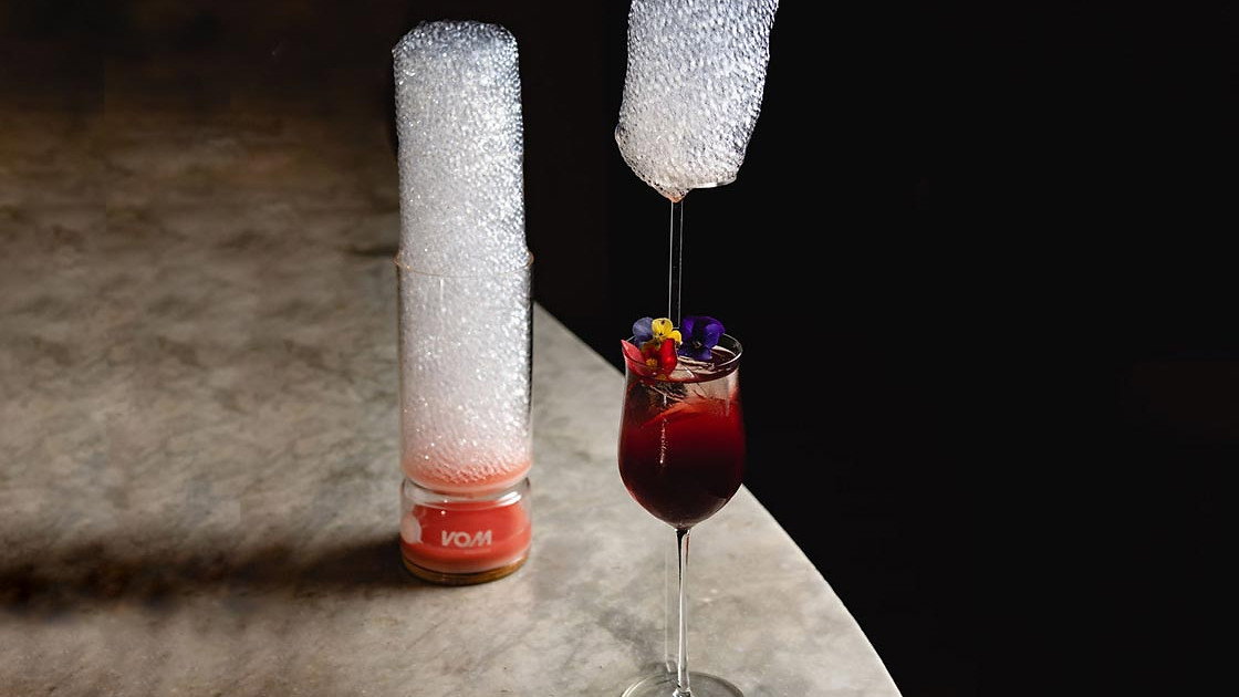 A cocktail with floral garnish next to a tall glass with foamy bubbles on a marble table. Dark background, elegant and artistic scene.