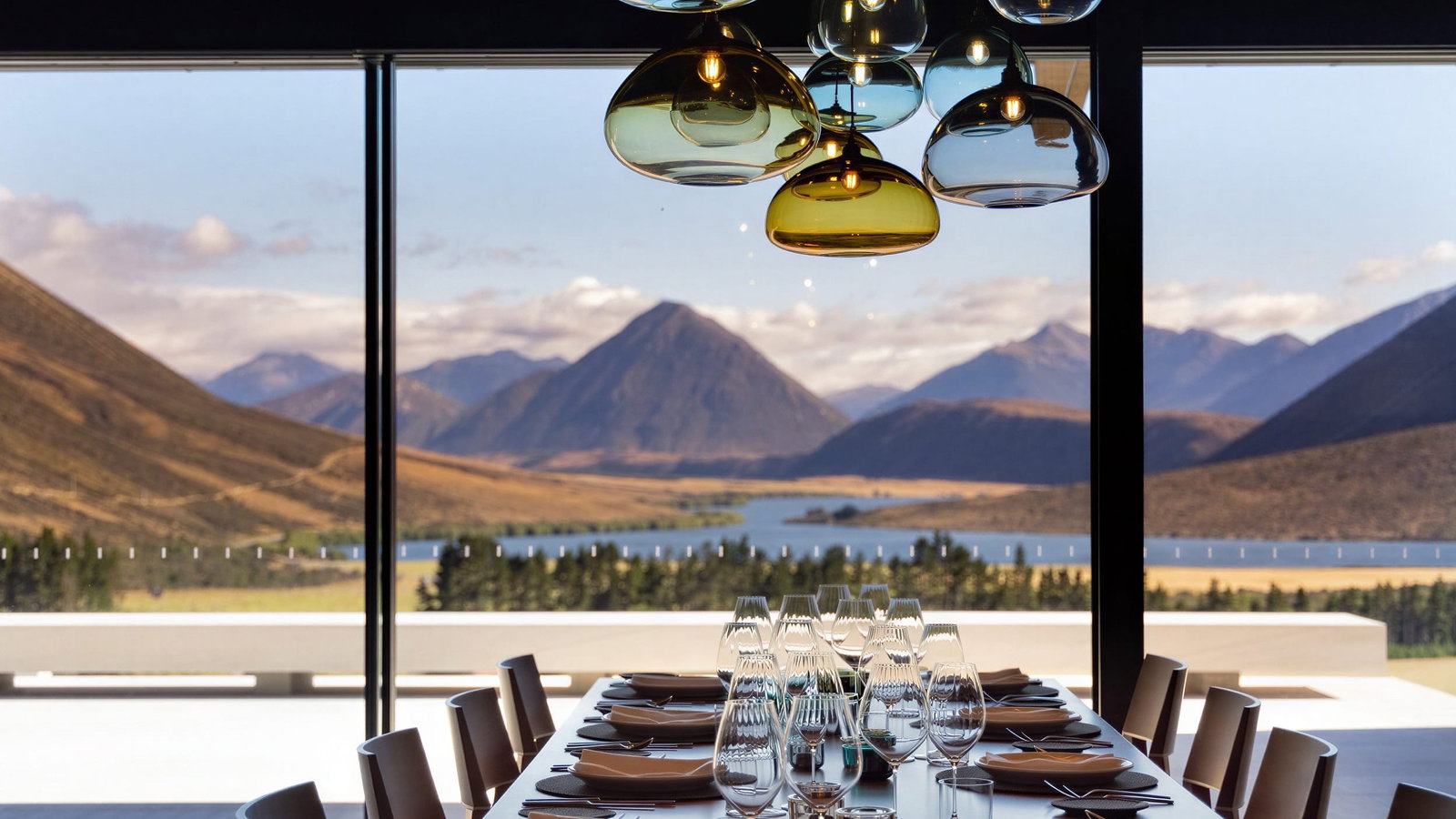 Dining table view at Flockhill Lodge overlooking mountains and a lake through large windows. Hanging colored lights create a cozy ambiance.