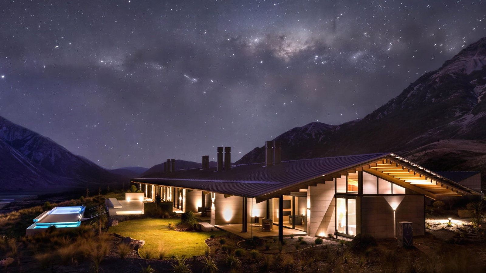 Flockhill Lodge exterior with warm lights under a starry sky, surrounded by mountains; a glowing pool and illuminated patio create a serene night scene.