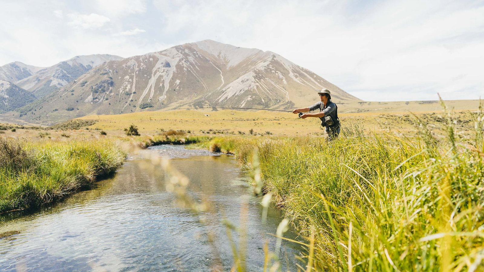 Person fly-fishing at Flockhill Lodge in a grassy field by a stream, with mountains in the background under a clear sky, creating a serene outdoor scene.