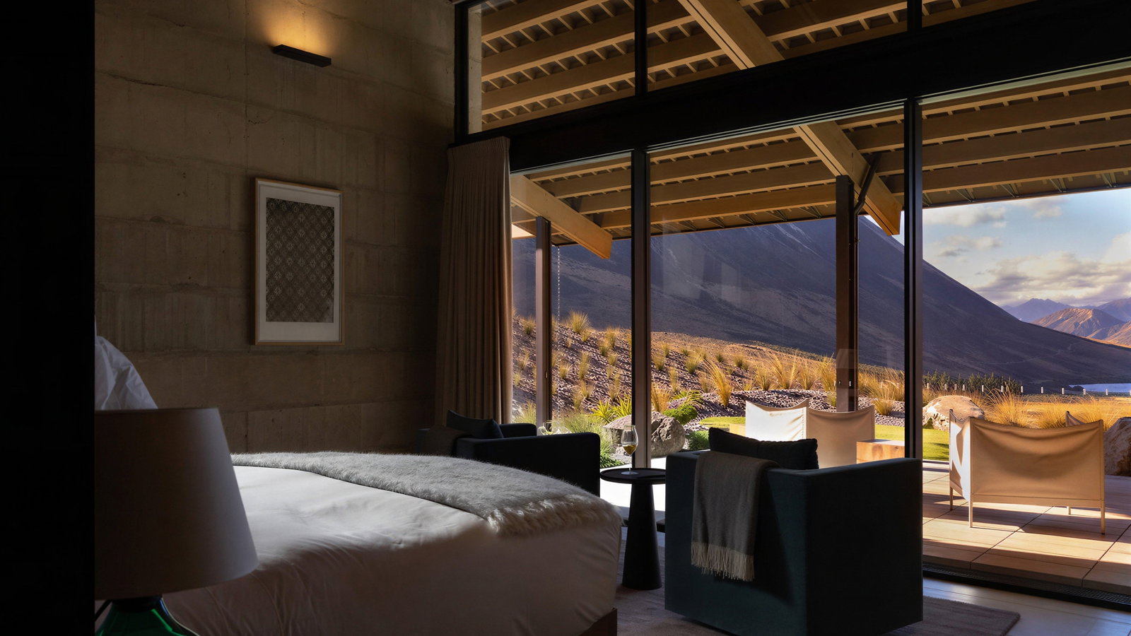 The Homestead bedroom at Flockhill Lodge with large windows overlooking mountains. Sunlit patio with two chairs. Neutral tones create a serene atmosphere.