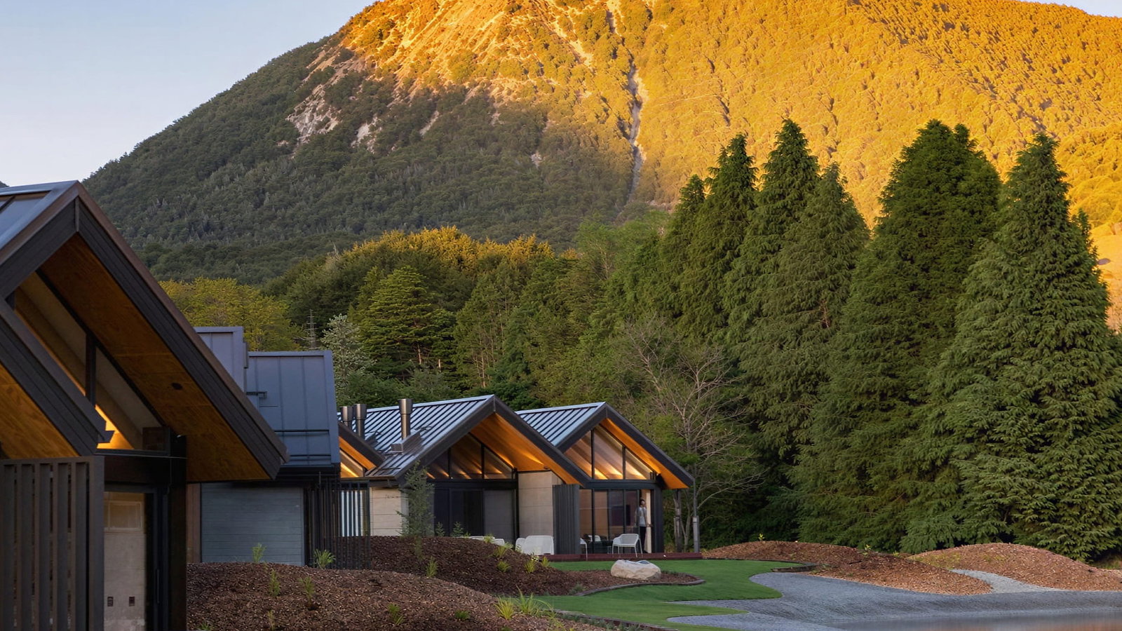 Modern villas at Flockhill Lodge with metal roofs sit near a serene pond, surrounded by lush trees and a sunlit mountain under a clear sky.