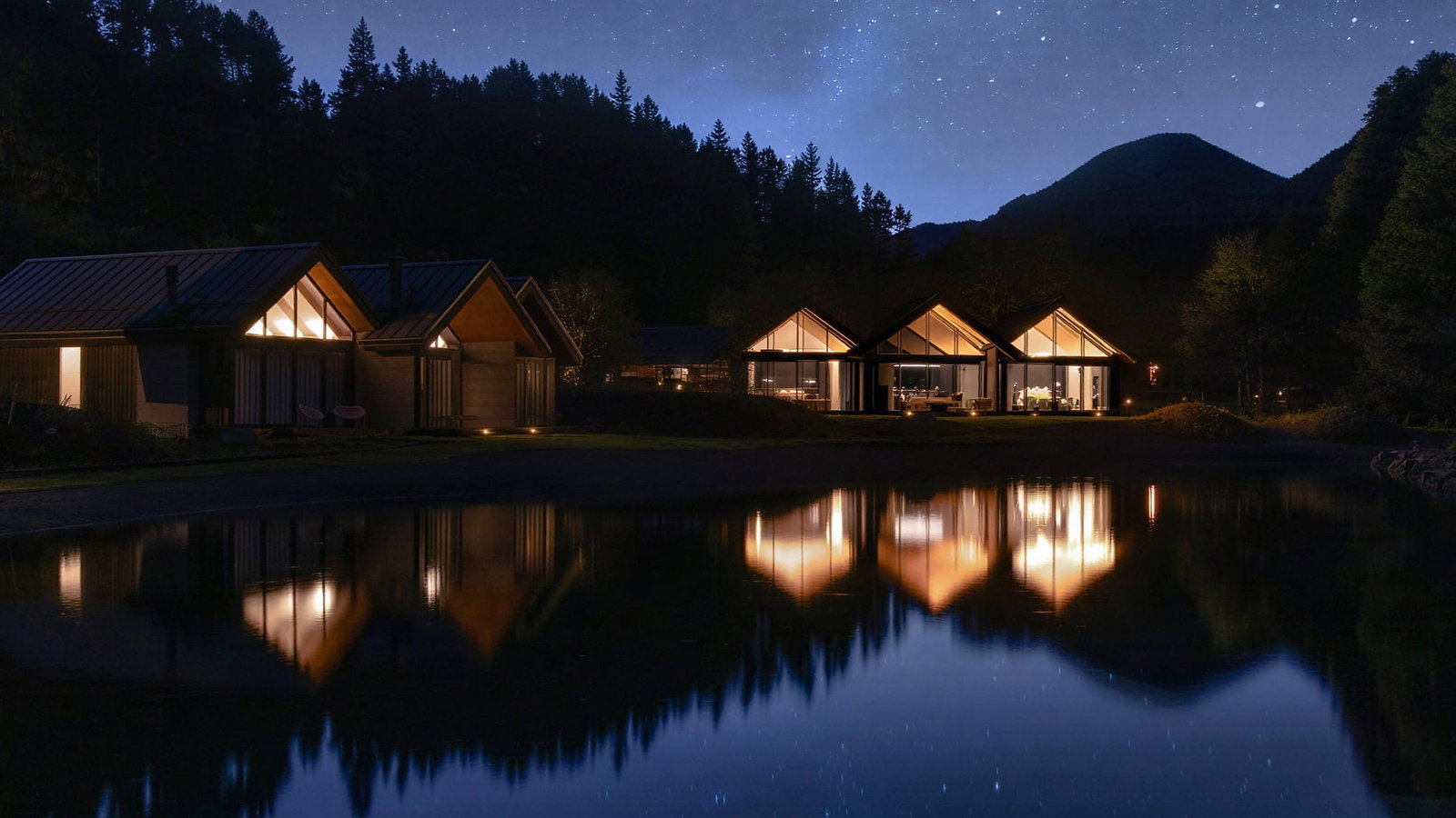Villas at Flockhill Lodge with lit windows reflect in a calm lake under a starry night sky, surrounded by silhouetted trees and mountains, creating a serene scene.