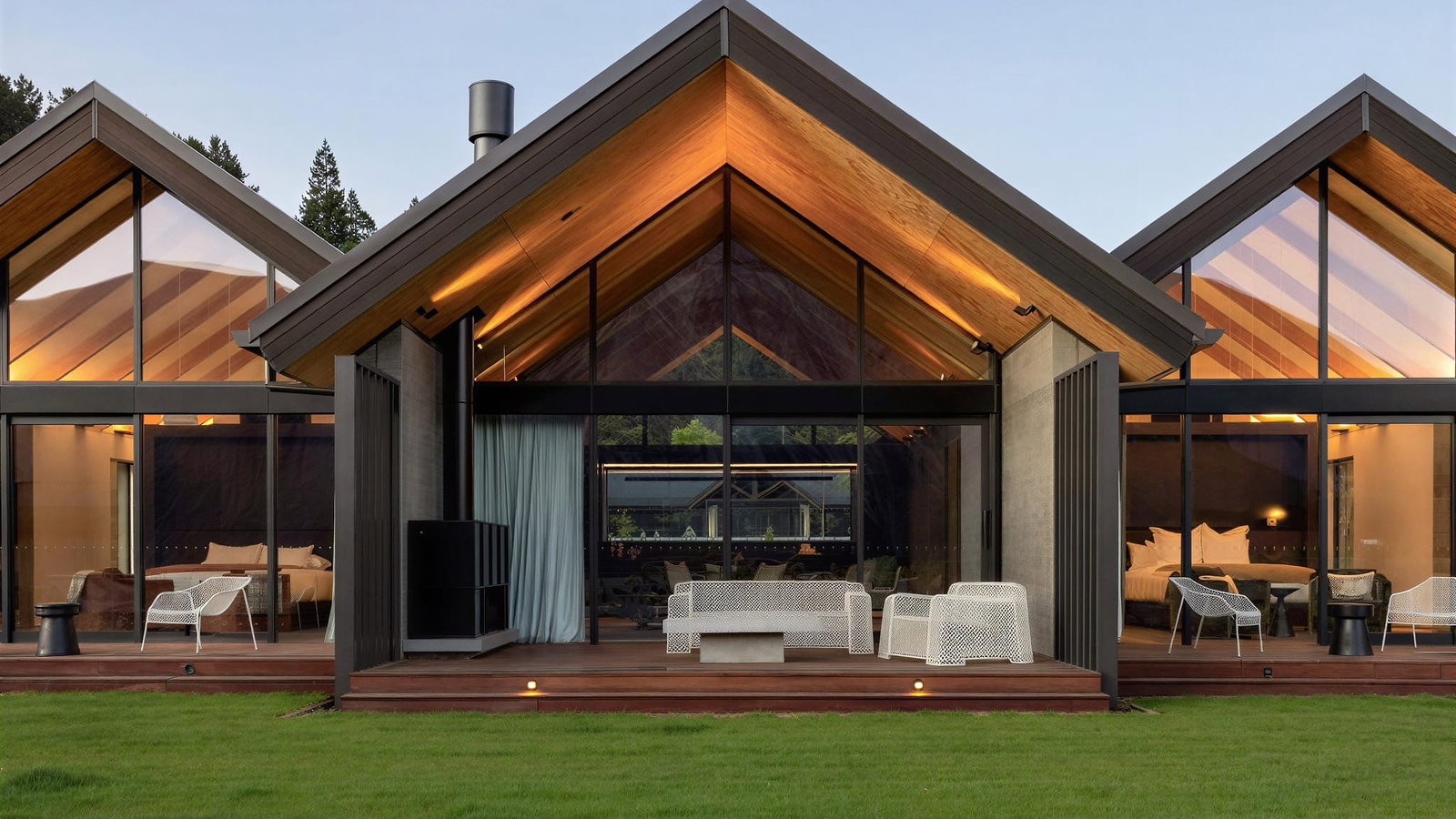 Modern villas at Flockhill Lodge with large glass windows, warm lighting, and triangular roofs. Patio features white chairs on wooden decking. Lush green lawn.