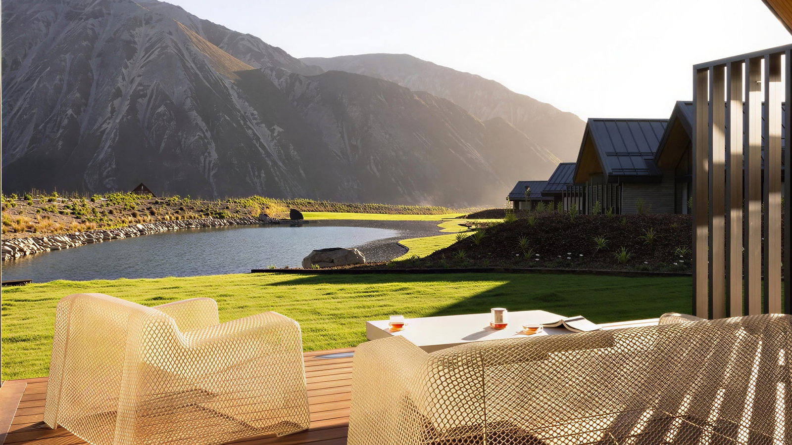 Outdoor deck view from a Villa at Flockhill Lodge with mesh chairs and table, two drinks, and a book. Overlooks grass, pond, and mountains under soft sunlight, modern cabins nearby.