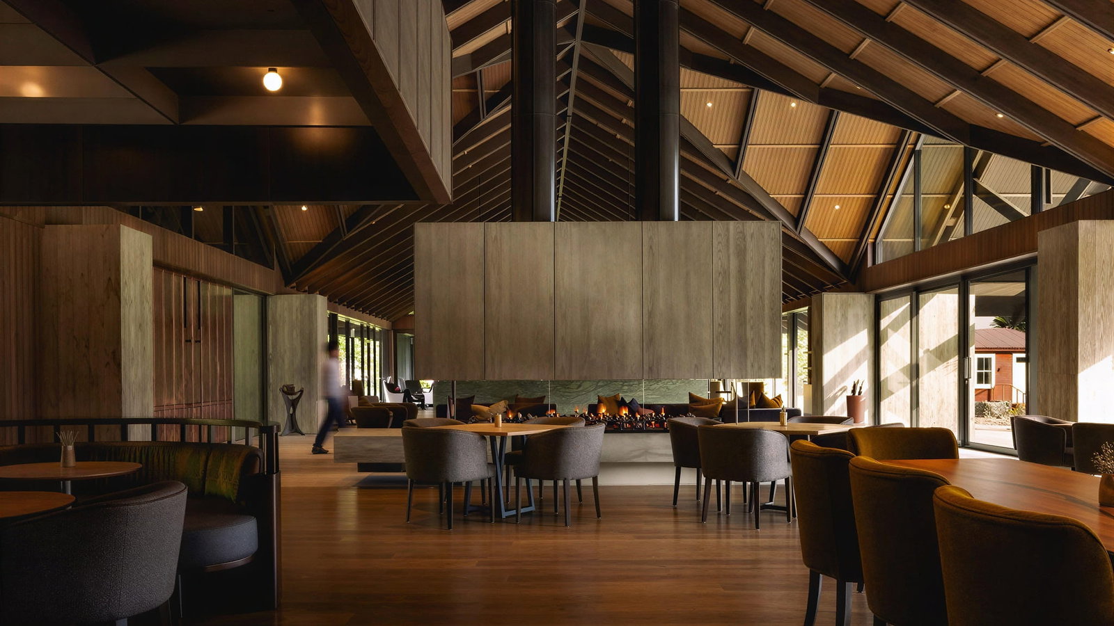 Spacious dining Sugarloaf restaurant dining room at Flockhill Lodge with wooden ceiling, central fireplace, dark chairs, and large windows. A person walks past, creating a serene atmosphere.
