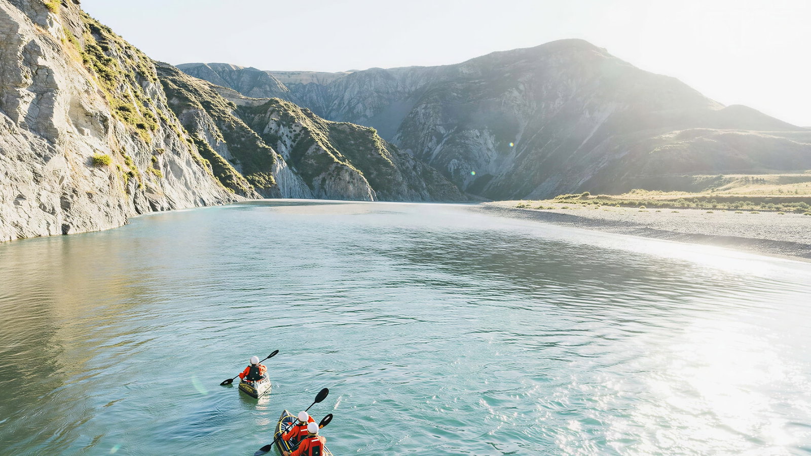 Kayakers at Flockhill Lodge in red gear paddle through a serene, sunlit river with rocky cliffs and mountains in the background. Peaceful and scenic setting.