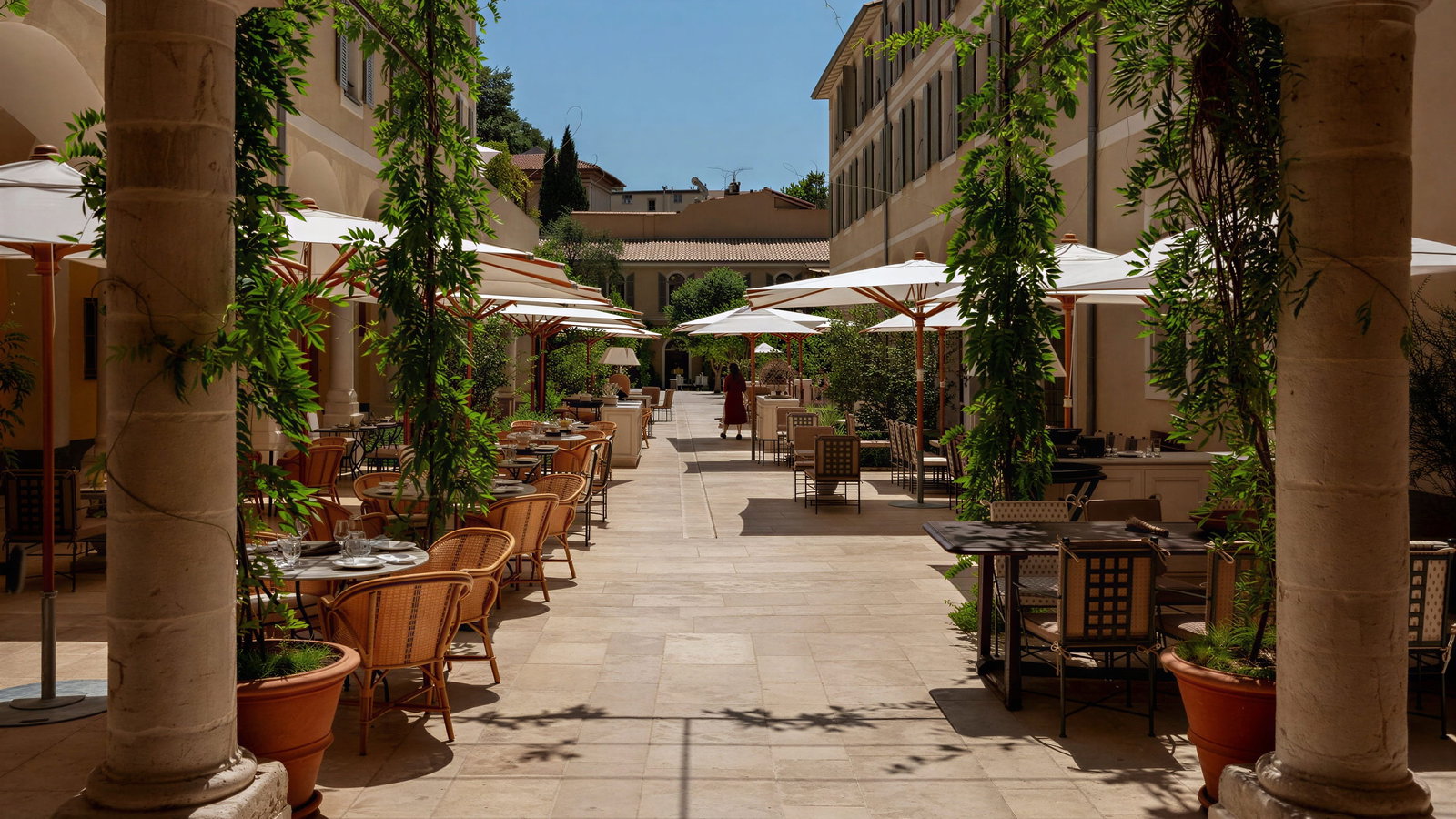 Sunlit outdoor courtyard at Hôtel du Couvent with empty wicker chairs and tables, white umbrellas, greenery, and columns. Beige walls and a clear blue sky. Peaceful mood.