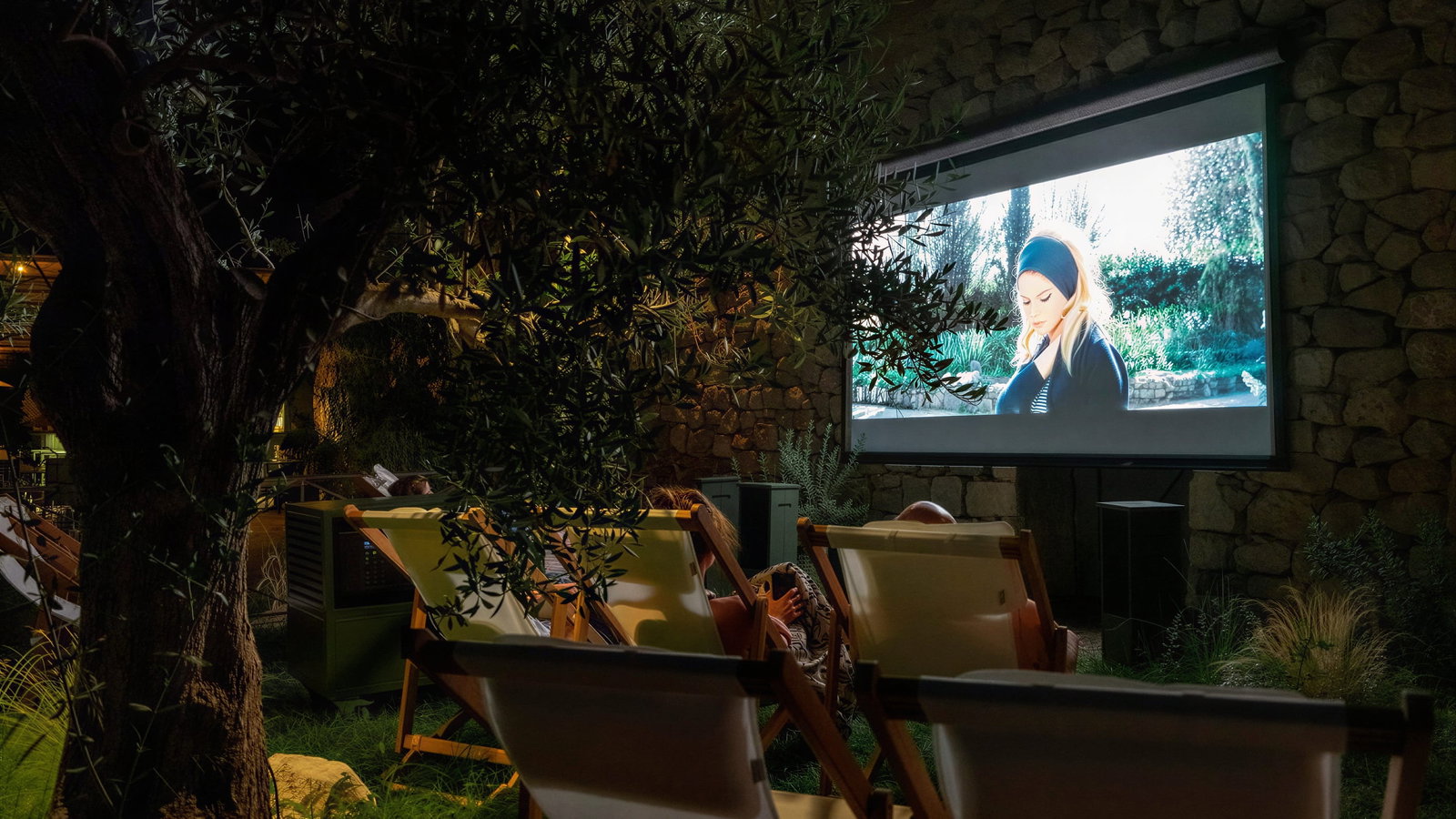 Outdoor movie screening at Hôtel du Couvent. People in lounge chairs watch on a large screen. The setting is a dimly lit garden with stone walls and greenery.