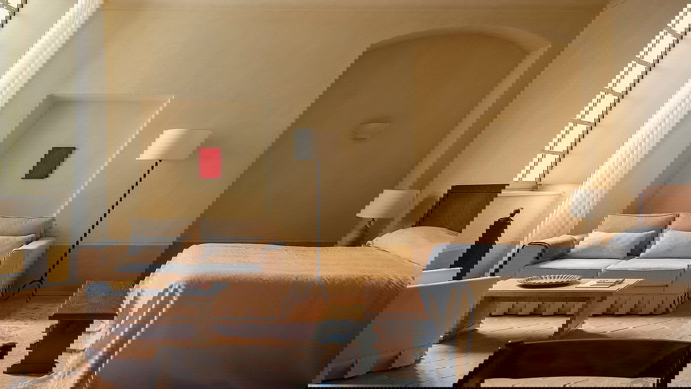 Suite du Noviciat room at Hôtel du Couvent with beige sofa, bed, and lamps. A small red painting hangs on the wall. Warm lighting creates a calm, inviting atmosphere.