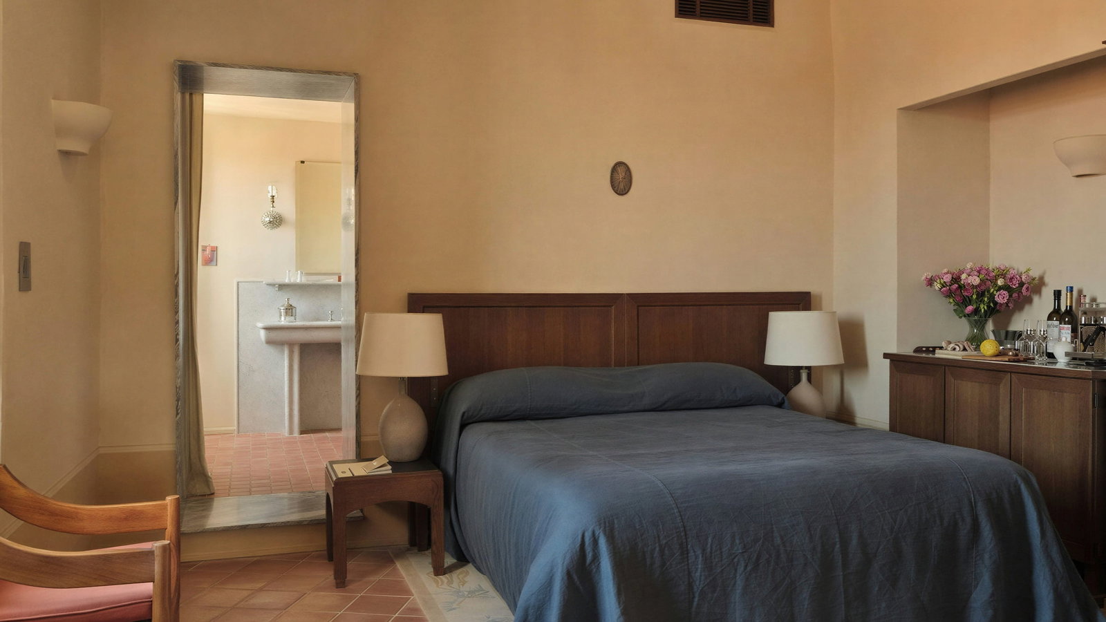 Colettines Room at Hôtel du Couvent with brown wood furniture, a bed with a blue cover, a mirror reflecting a bathroom, flowers on a dresser, and soft lighting.