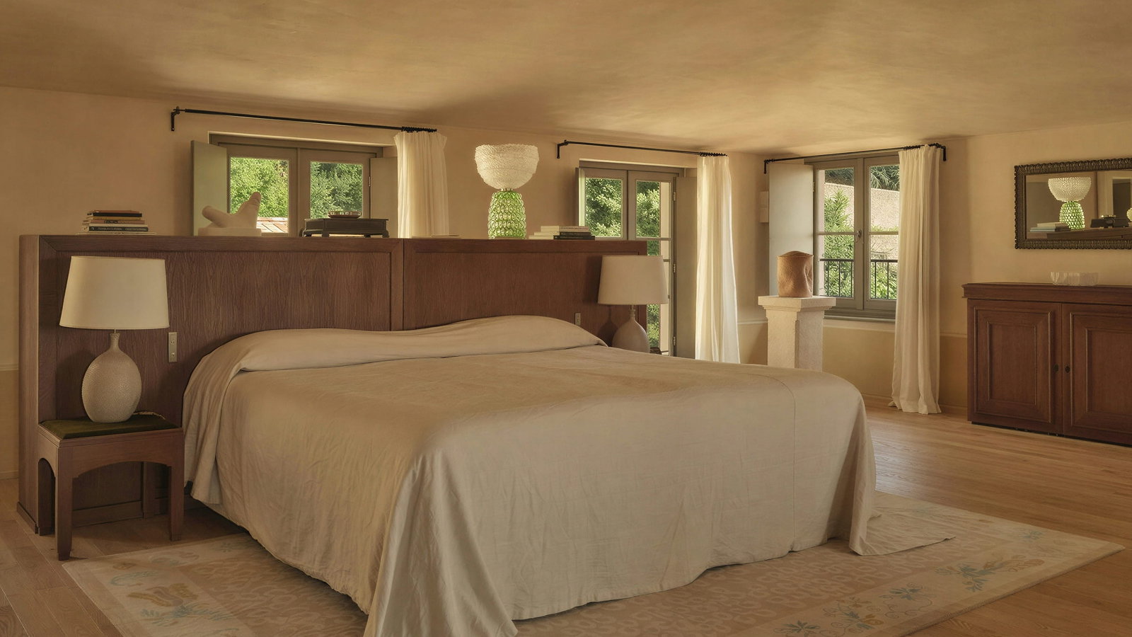 Suite la tour master bedroom at Hôtel du Couvent with a beige bed, wooden headboard, and lamps. Windows with curtains let in natural light. Calm and inviting atmosphere.