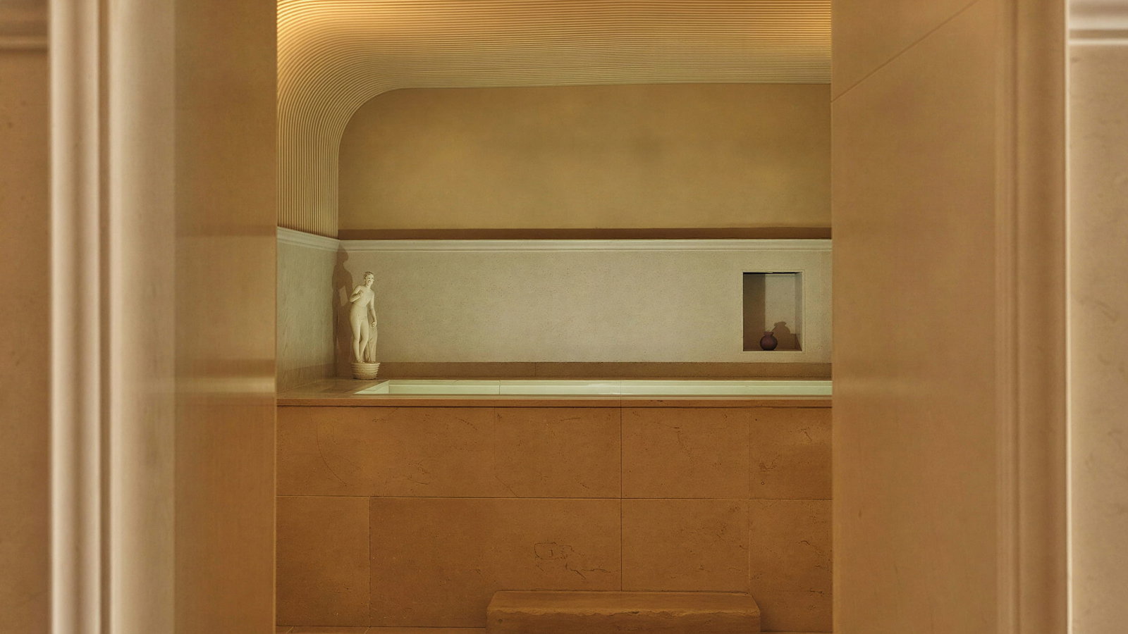 Roman Baths at Hôtel du Couvent with beige walls and a small statue. Soft lighting creates a warm atmosphere. A niche holds a small vase for decoration.
