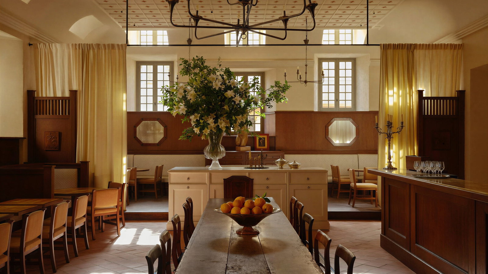 Hôtel du Couvent restaurant dining room with long table, vase of flowers, and bowl of oranges. Warm lighting, wooden furniture, and yellow curtains create a cozy ambiance.
