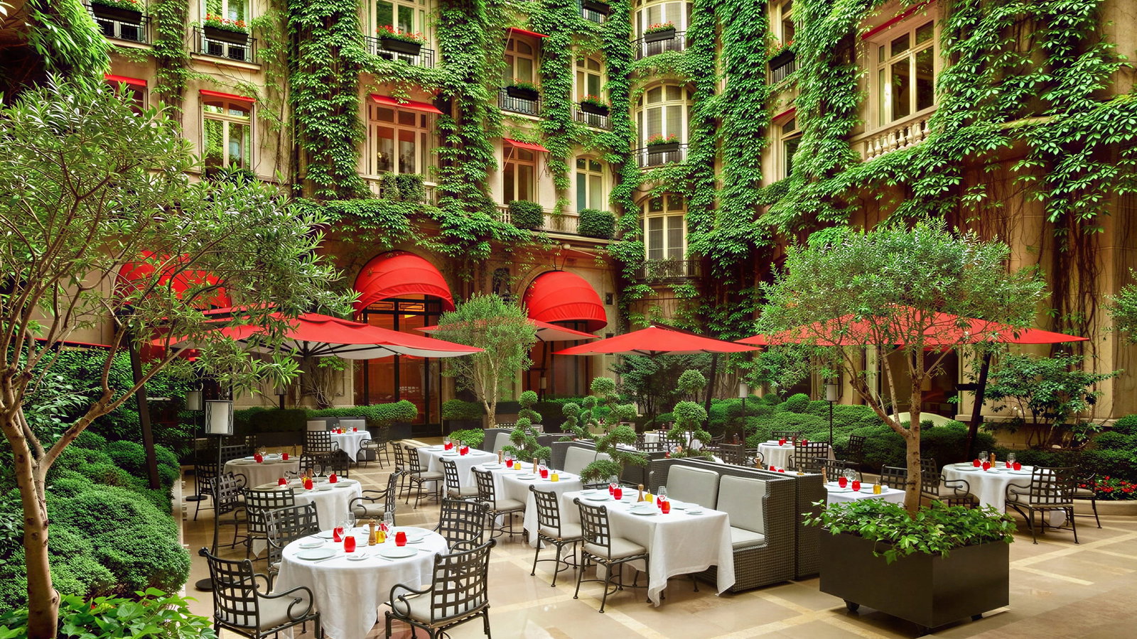 Outdoor courtyard café at Hôtel Plaza Athénée with white tablecloths and red cups under red umbrellas. Green ivy covers the building, creating a lush, inviting setting.
