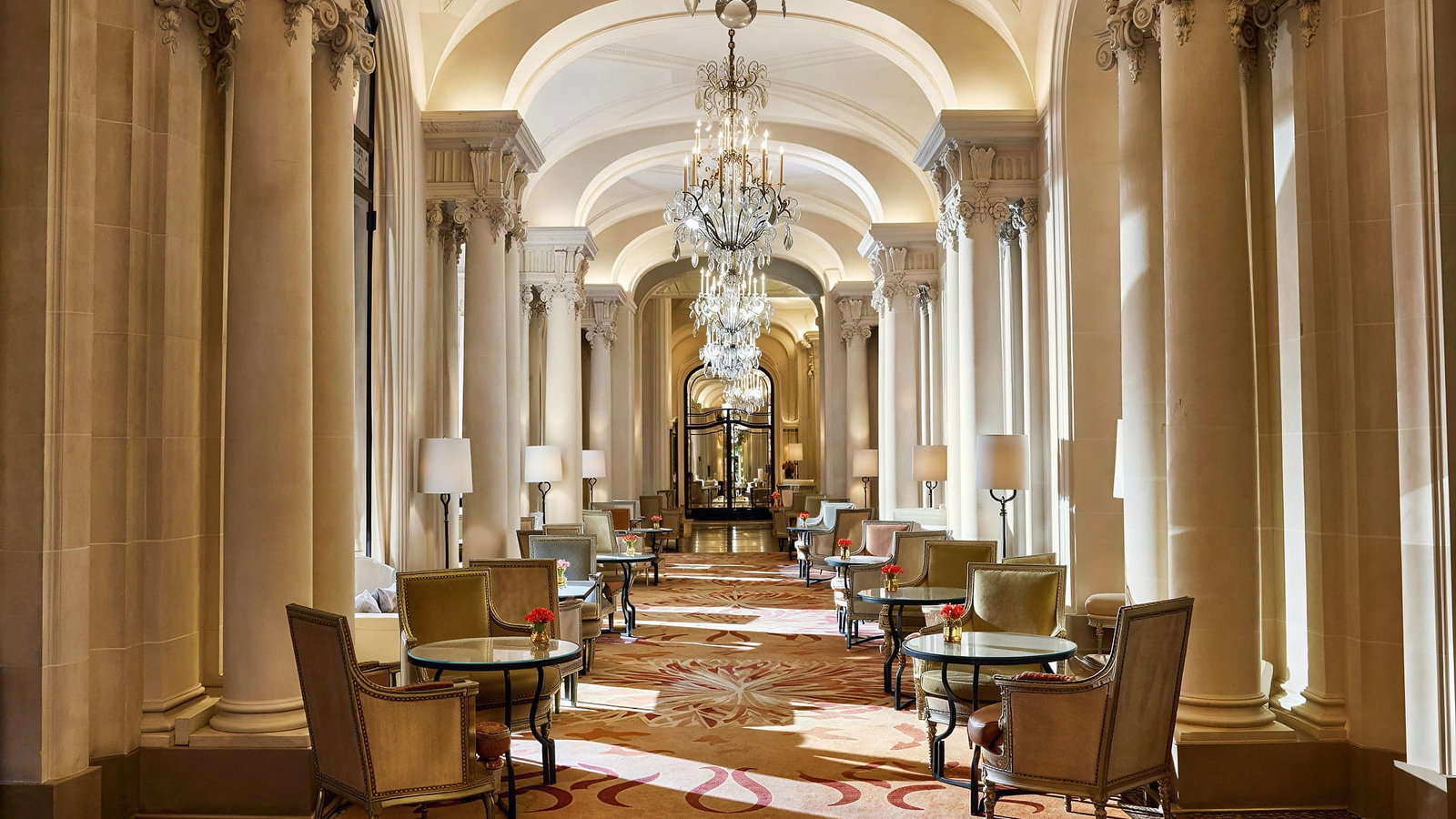 Elegant hotel hallway La Galerie at Hôtel Plaza Athénée with ornate pillars, chandeliers, and plush chairs around glass tables. Warm lighting and decorative carpet enhance luxury.