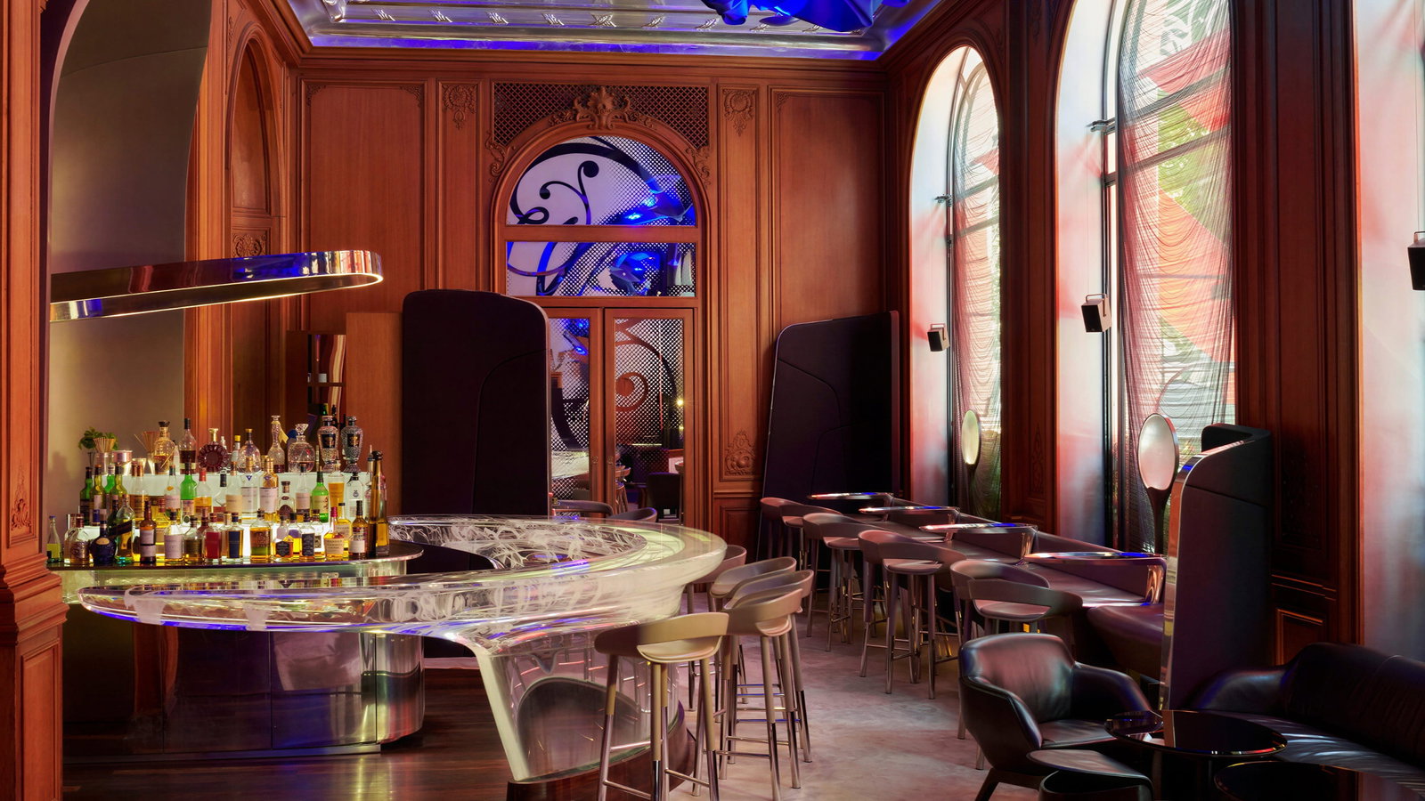 Sophisticated Le Bar interior at Hôtel Plaza Athénée with a sleek, translucent bar counter, stools, and plush seating. Blue ceiling art, wooden walls, and arched windows.