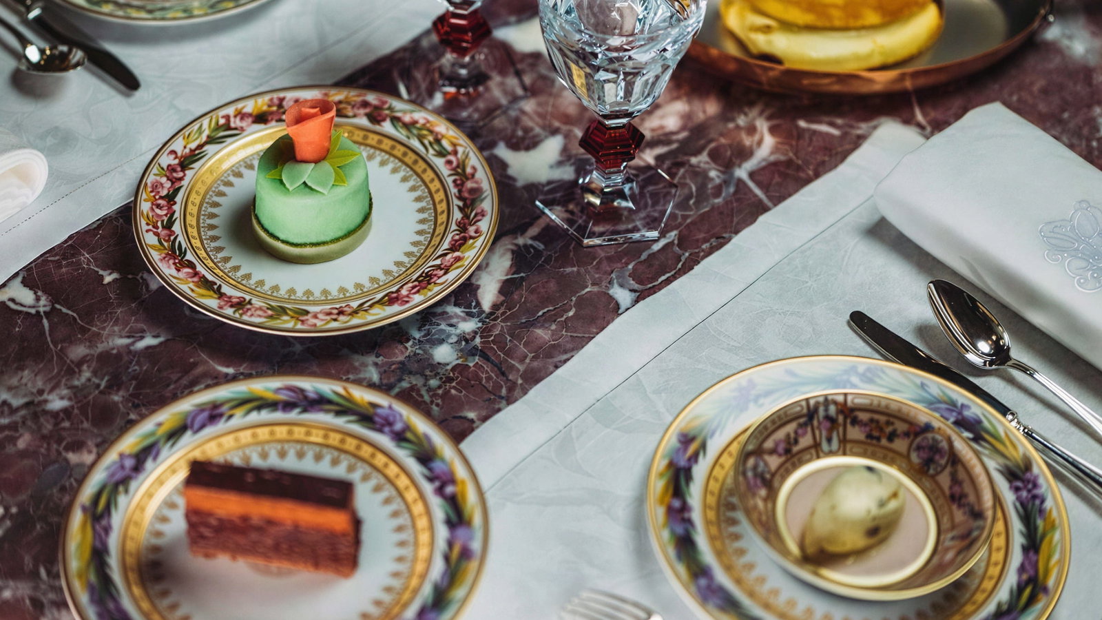 Elegant table setting at Jean Imbert at Hôtel Plaza Athénée with ornate plates, colorful desserts, crystal glasses, and folded napkins on a marbled tablecloth. Sophisticated mood.