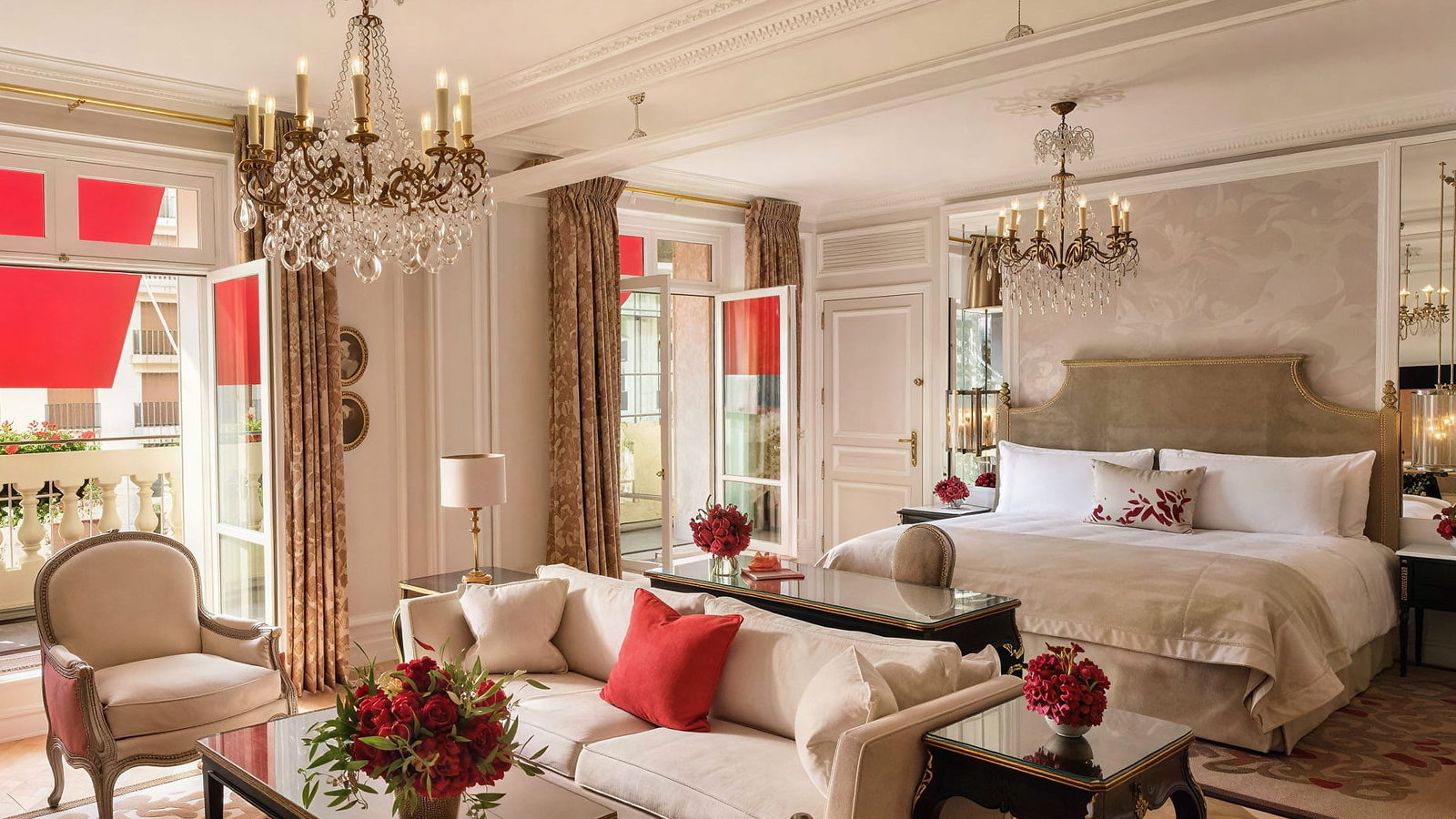Elegant Junior Suite Avenue Montaigne View room at Hôtel Plaza Athénée with a plush bed, chandelier, beige and red decor, and a sunlit balcony. Cozy seating area with floral accents.