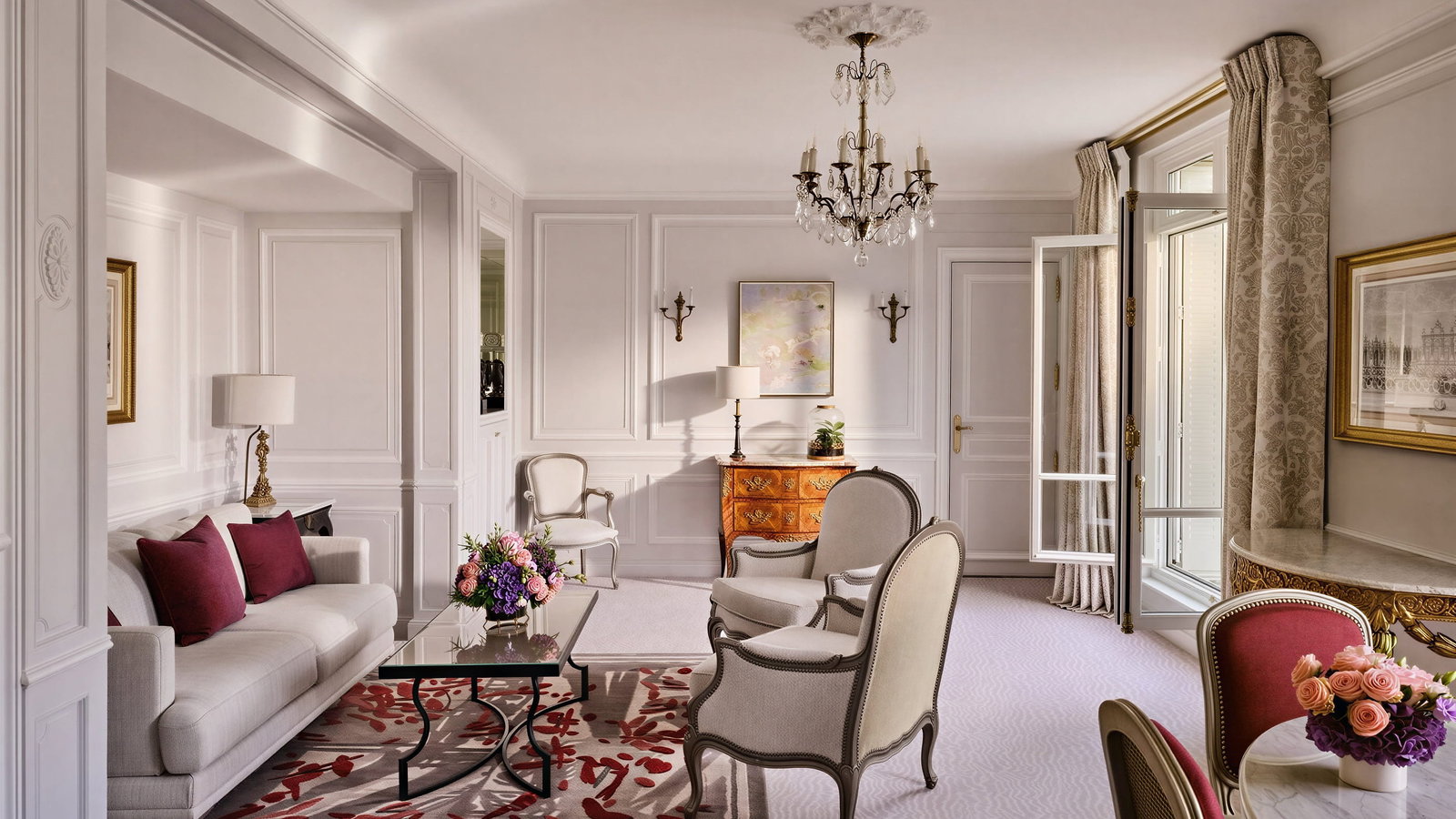Elegant prestige suite living room at Hôtel Plaza Athénée with white walls, chandelier, red-accented rug, white sofa, armchairs, and flowers on a table; large windows with curtains.