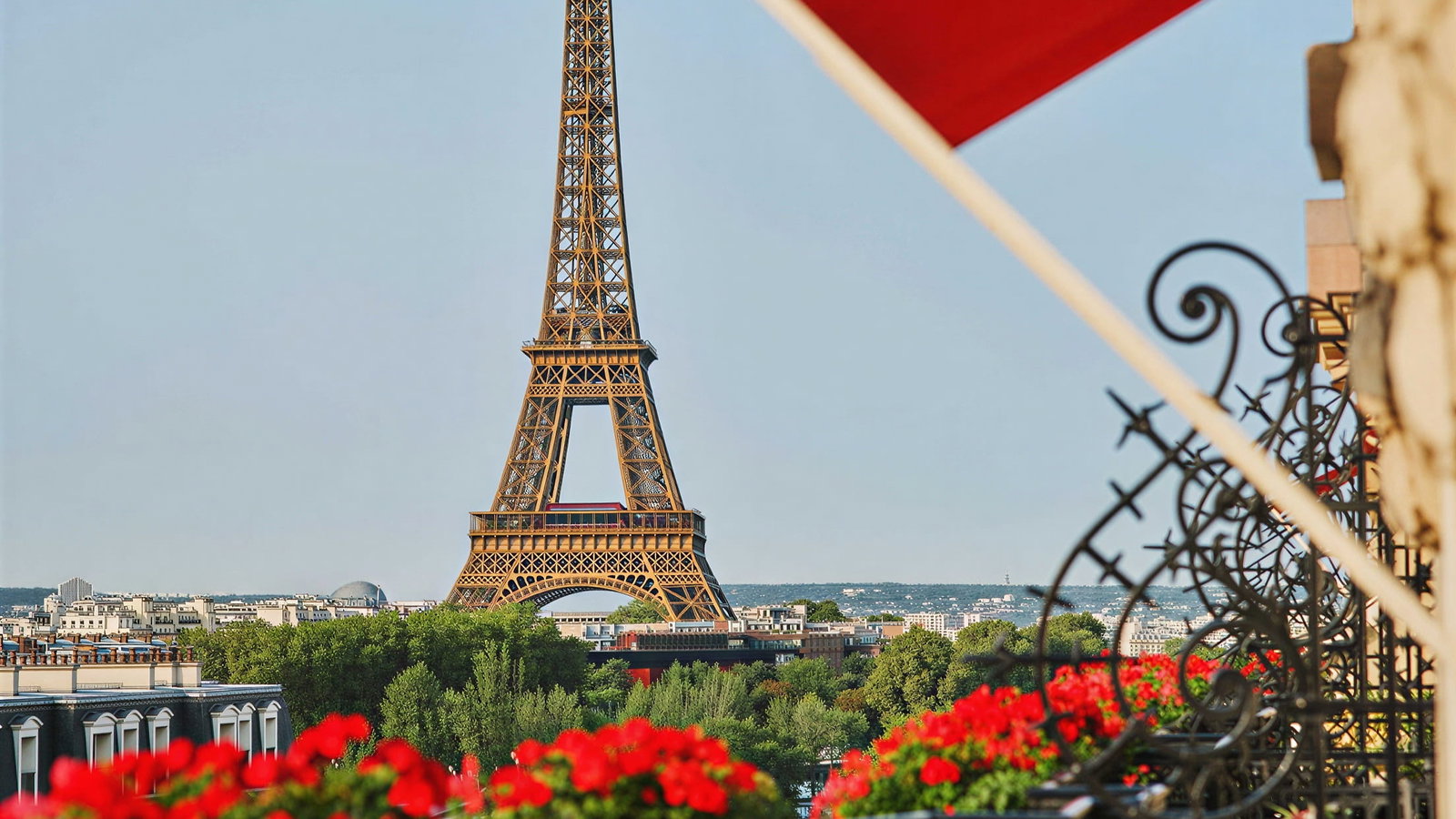 Signature Eiffel Suite Balcony Eiffel Tower view at Hôtel Plaza Athénée with red flowers. Breakfast is on a table with a white cloth and red chairs. Bright, sunny mood.