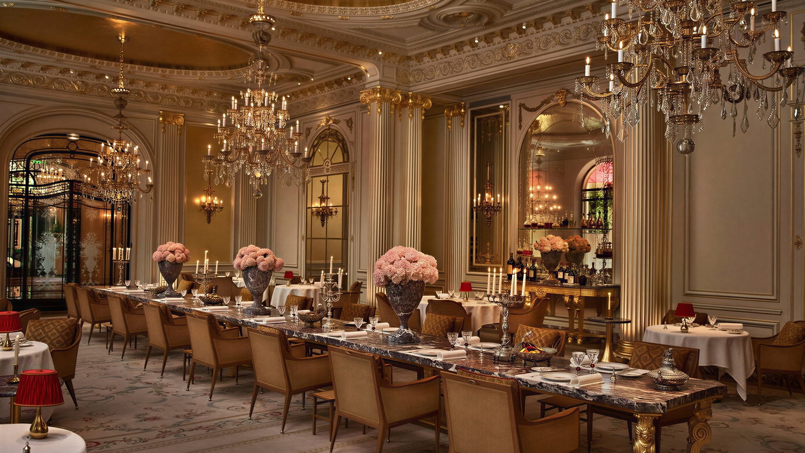 Elegant Jean Imbert dining room at Hôtel Plaza Athénée with chandeliers, ornate ceiling, and floral decor. Long tables set with candles and red lamps create a luxurious ambiance.