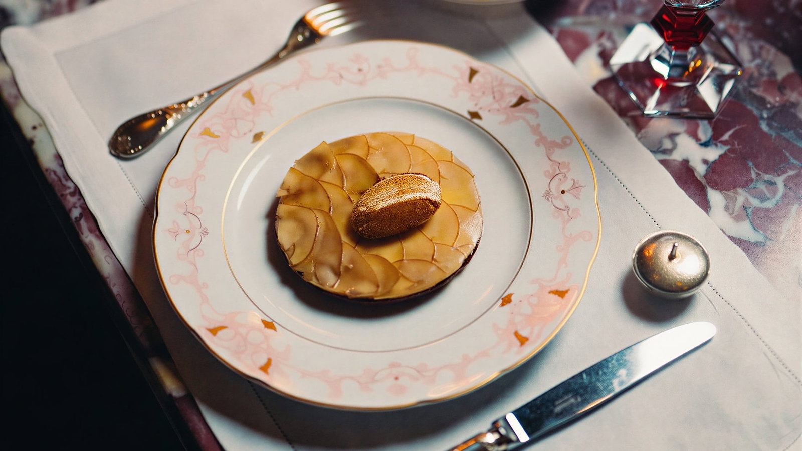 Elegant setting with a plated La Tarte aux Cepes dessert on floral china at Hôtel Plaza Athénée, a colorful salad in a bowl, a glass of water, and a silver knife and fork on a marble table.
