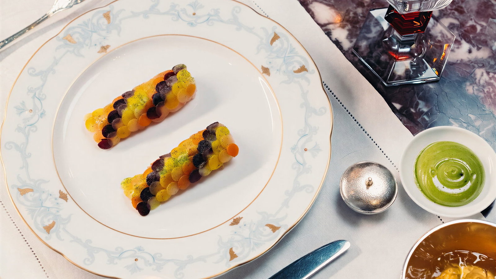 Elegant dining setup of Les Langoustines à la Parisienne from Jean Imbert at Hôtel Plaza Athénée on a patterned plate, a glass of water, sauces, and dessert on a marbled table.