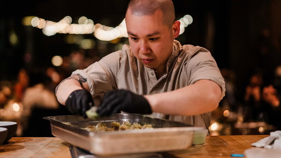 Chef in beige shirt and black gloves prepares food in a metal tray on a wooden table. Blurred restaurant setting with warm lights.