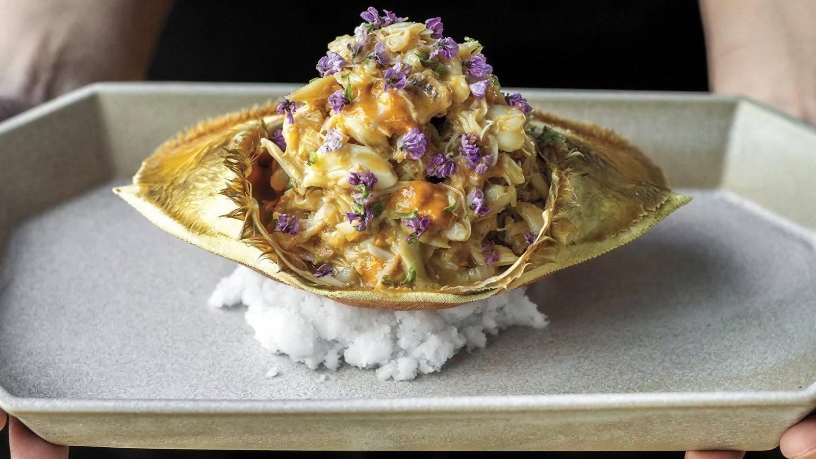 A seafood dish with a crab shell filled with salad and purple flowers on a bed of salt, presented on a beige tray by a person’s hands.