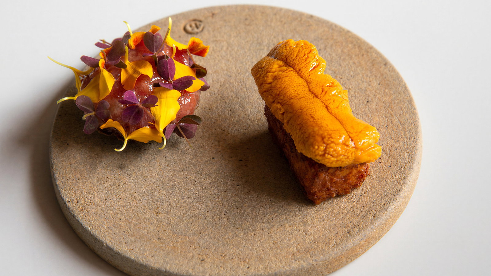 Gourmet dish on a beige stone plate: one piece with yellow uni on brown base, another garnished with yellow petals and purple leaves.