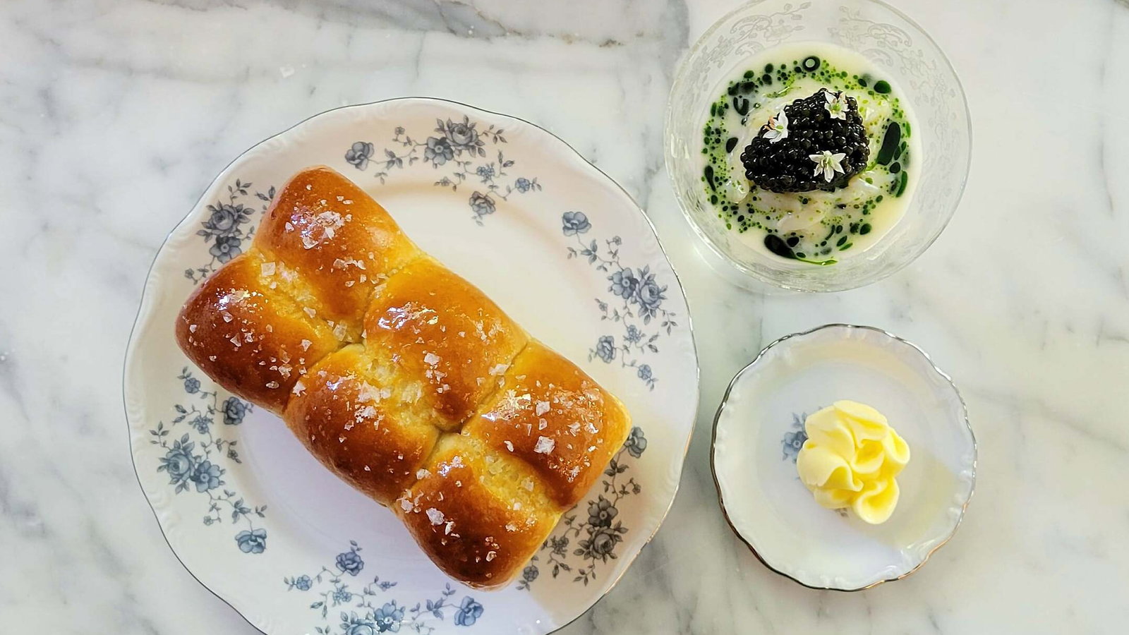 Bread topped with salt on a floral plate, caviar dish with green garnish, and butter on marble surface. Elegant dining setup.
