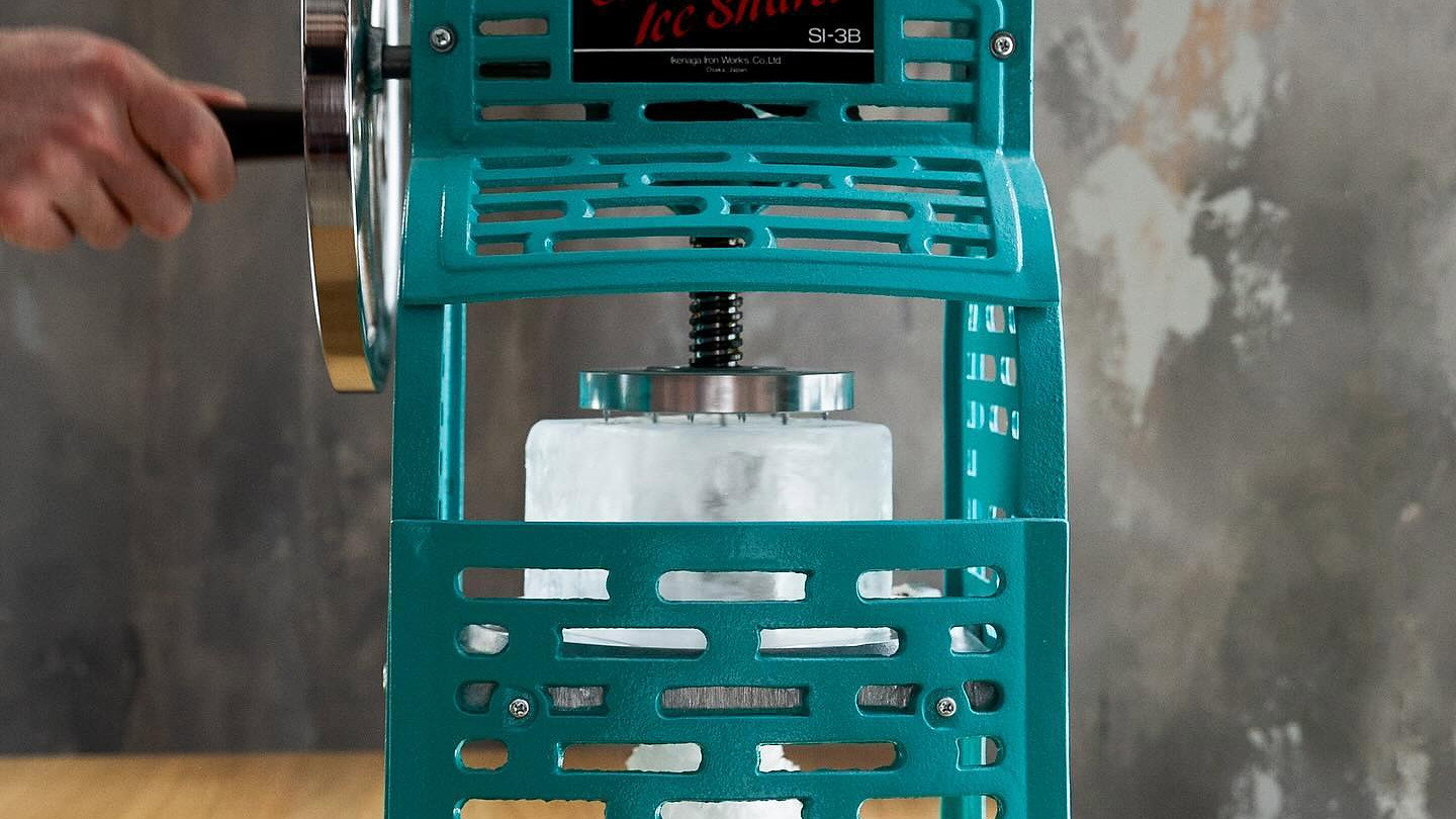 A teal Swan ice shaver on a wooden table shaves ice into a glass below. Background is gray and textured. A hand turns the handle.