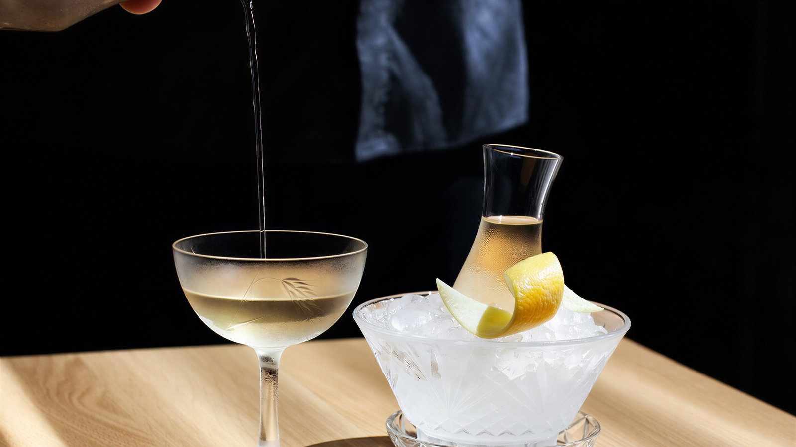 A hand pours a clear liquid into a frosted glass on a wooden table. Beside it, a carafe with a lemon twist sits in a bowl of ice.