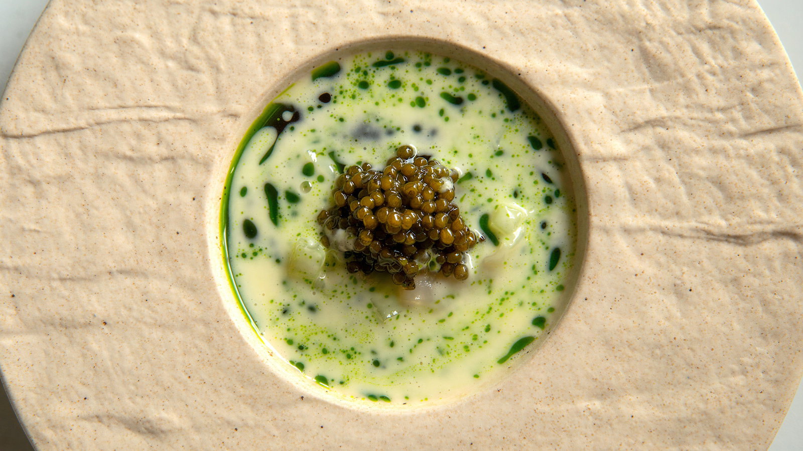 Caviar atop creamy soup with green herbs, presented in a textured beige bowl. The setting is minimalist with a sophisticated feel.