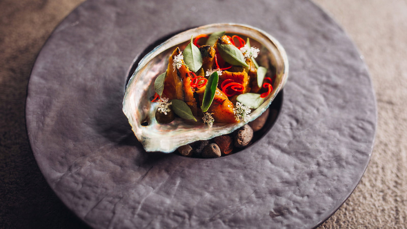 Gourmet dish in a shell on a textured, gray plate. Garnished with red curls, green leaves, and white flowers, creating an artistic presentation.