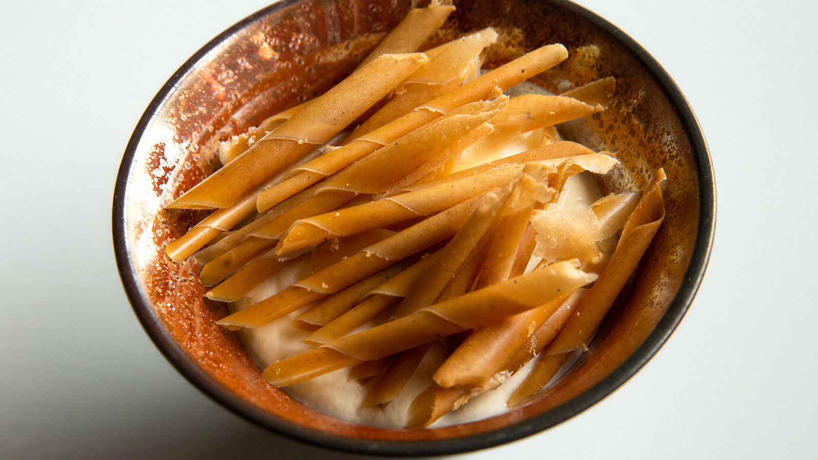 Bowl of ice cream topped with thin, twisted caramel sticks. The bowl has a rustic brown glaze, set against a plain white background.