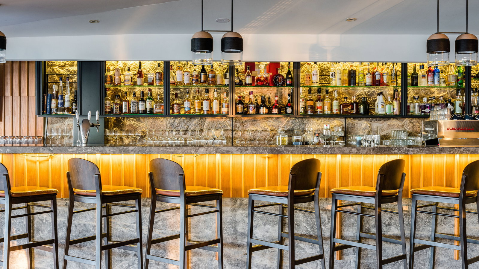 Salt Water Restaurant Bar at Lizard Island Resort with six wooden stools, a stone counter, illuminated shelves stocked with liquor bottles, and warm lighting.