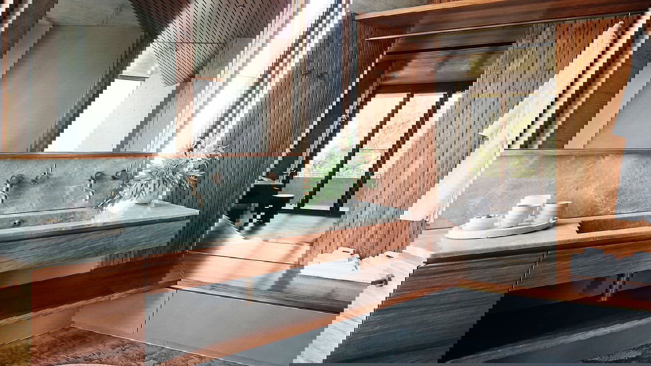 Modern bathroom at The House on Lizard Island Resort with wooden walls, green stone sink, and bronze fixtures. White robes hang neatly. Large window offers natural light. Calm atmosphere.