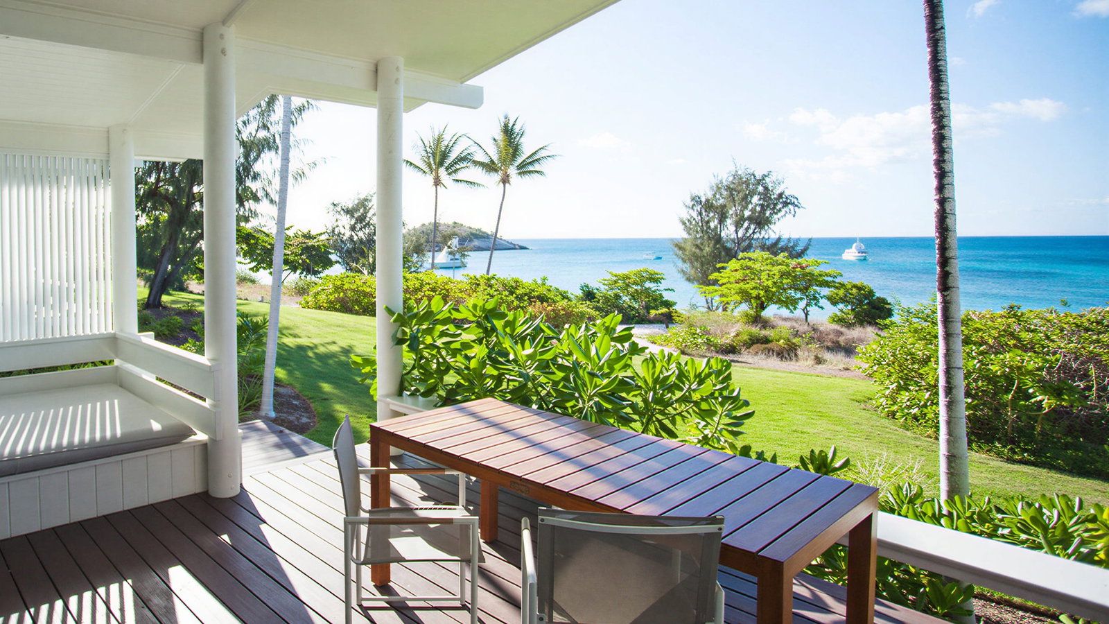 Oceanfront veranda with wooden table, chairs, overlooking lush garden and blue sea. Palm trees sway under a clear sky, tranquil mood.