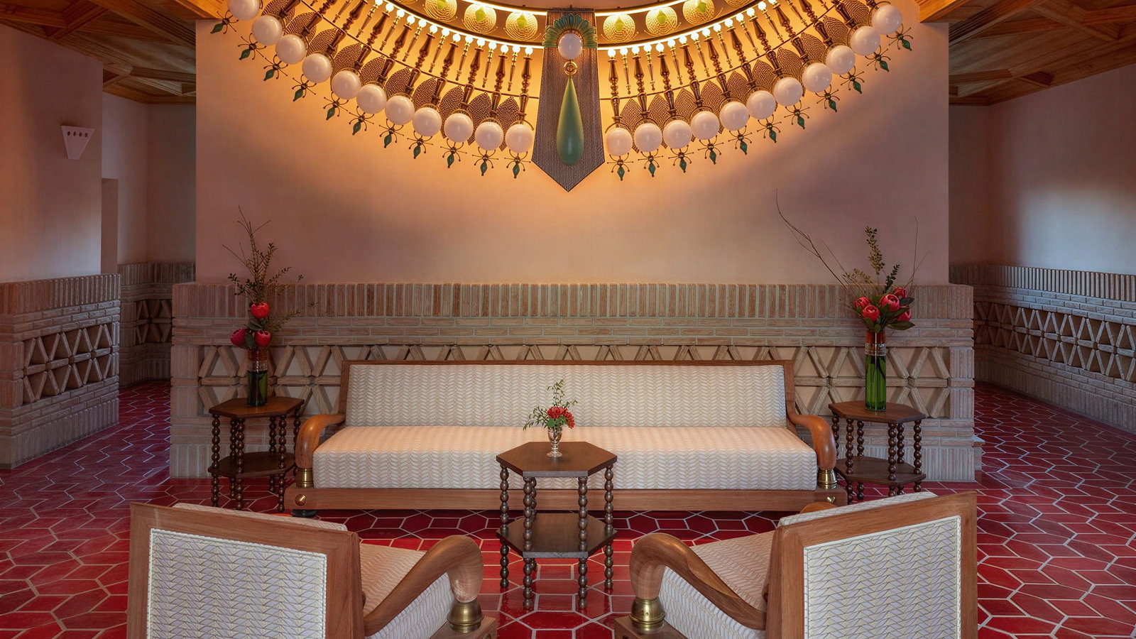 Vermelho Melides reception lobby with red hexagonal tiles, beige sofa, and chairs. Floral arrangements on tables. Illuminated wall decor adds warmth.