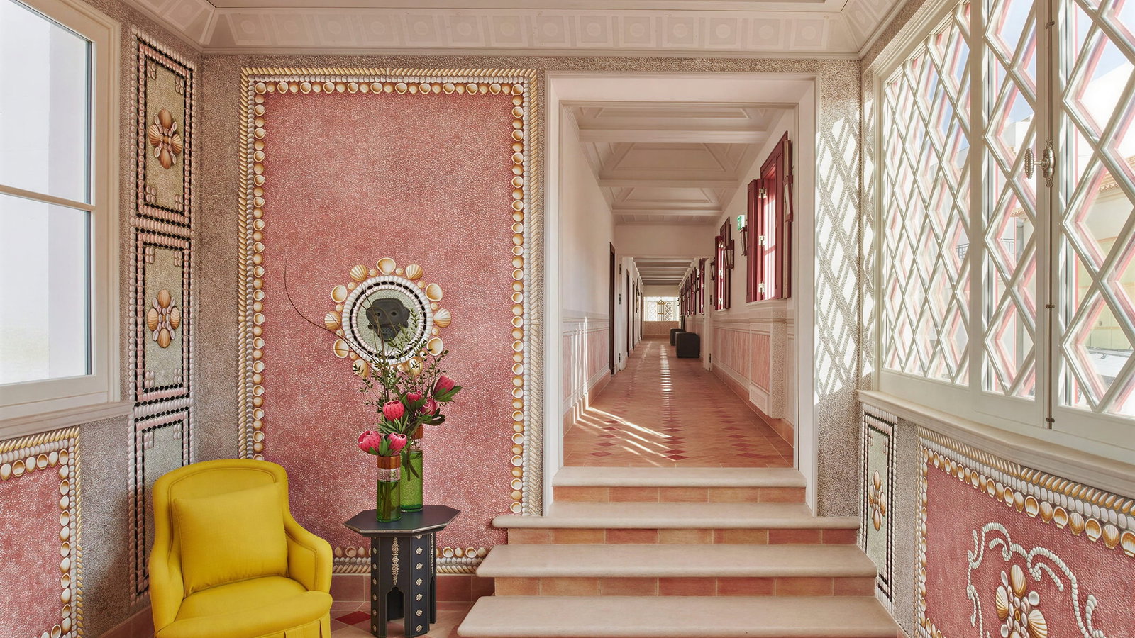 Ornate hallway at Vermelho Melides with pink walls, floral patterns, and a yellow armchair. Bright light filters through latticed windows. Vase with pink flowers.