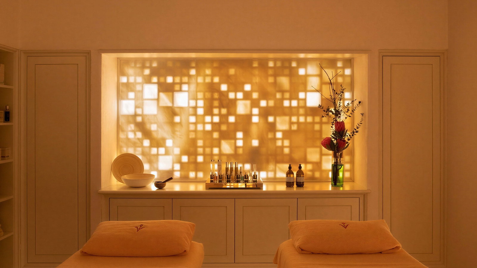 Two massage tables at Vermelho Melides Spa with orange linens face a backlit panel of geometric patterns. Vases, bowls, and bottles decorate the calm spa setting.